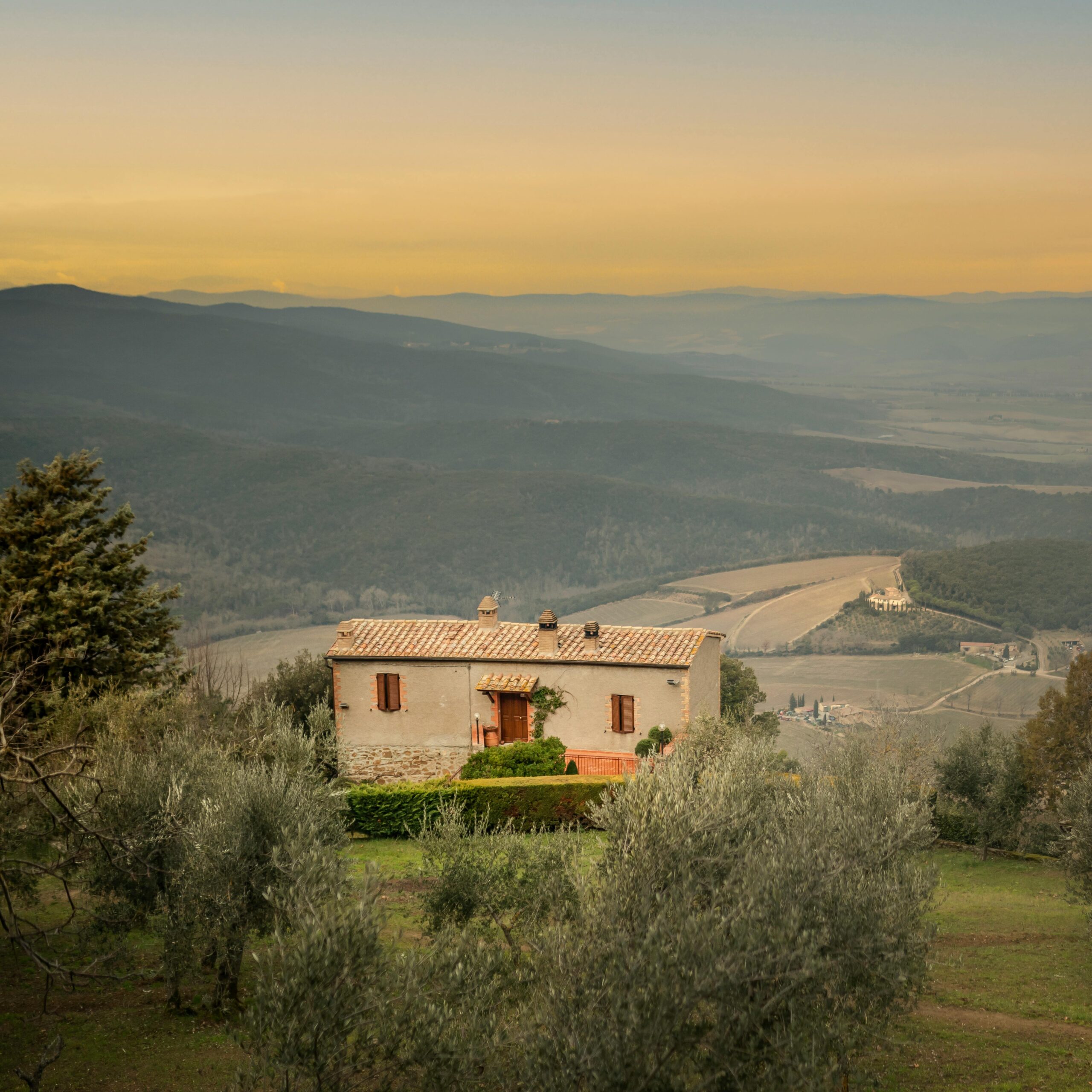 A serene view of a rustic house surrounded by rolling hills in the Tuscan countryside at sunset.